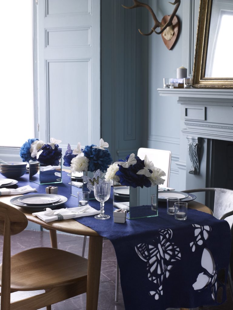 Dark navy blue dining room with fresh flowers centrepiece mirrors and butterflies with velvet table runner with butterflies and retro wooden chairs and table by Interior Stylist Maxine Brady