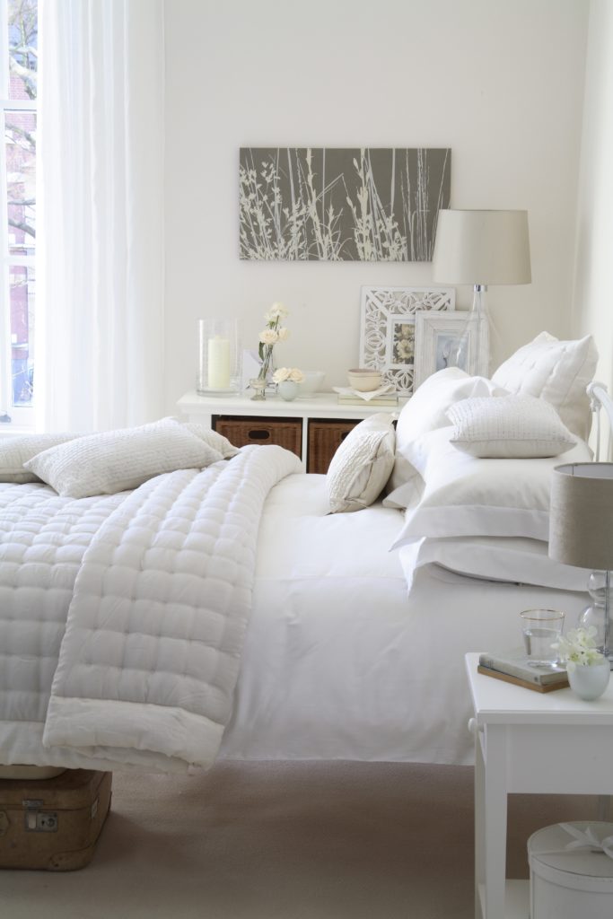 White bedroom with velvet quilt and scatter cushions shabby chic styling, vintgae suitcase with rattan storage by Interior Stylist Maxine Brady