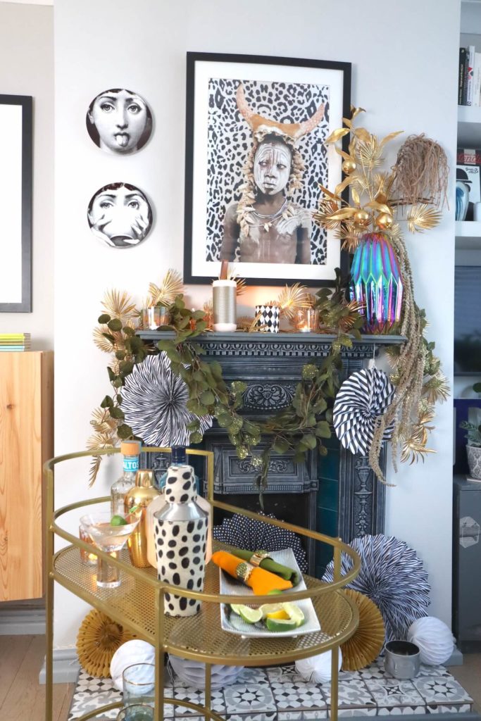 Fireplace & mantelpiece dressed with garland and paper decorations with cocktail gold trolley. black and white art. 7 cool Zoom Party Styling Ideas